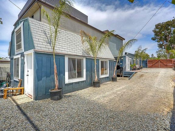 Front three-quarter view of the two blue board-and-batten studio cottages with a mansard-style roof, queen palms in pots along the gravel drive, and a wood gate at the rear of the parcel