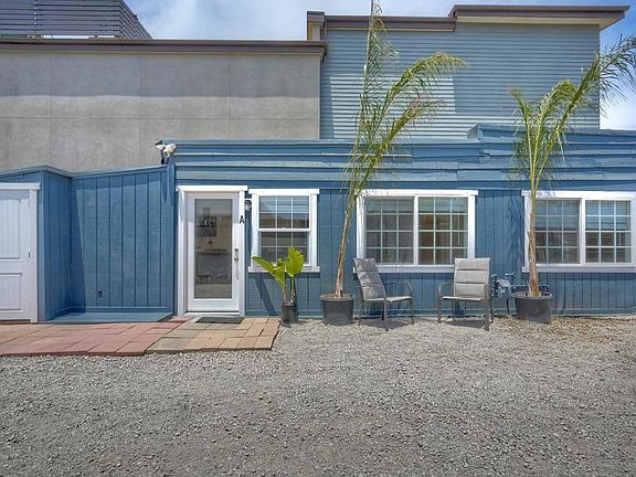 Rear-facing view of the blue studio cottages with a paver patio, two outdoor chairs, palms in pots, and a row of windows along the rear of one unit